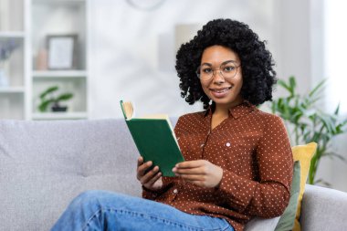Kanepede oturan, elinde bir kitapla rahatlayan ve kameraya bakıp gülümseyen Afro-Amerikan bir kadının portresi..