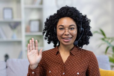 Kameranın önünde oturan ve selamlaşarak el sallayan, kulaklık takan gülümseyen Afro-Amerikan bir kadının yakın plan portresi..