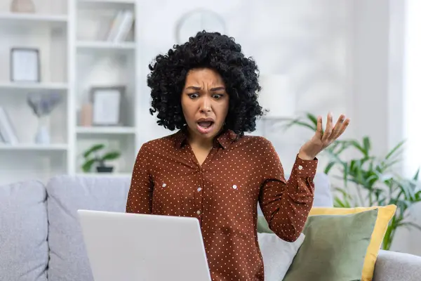 Angry young African American woman sitting on the couch at home and looking at the laptop screen with her hands in frustration.