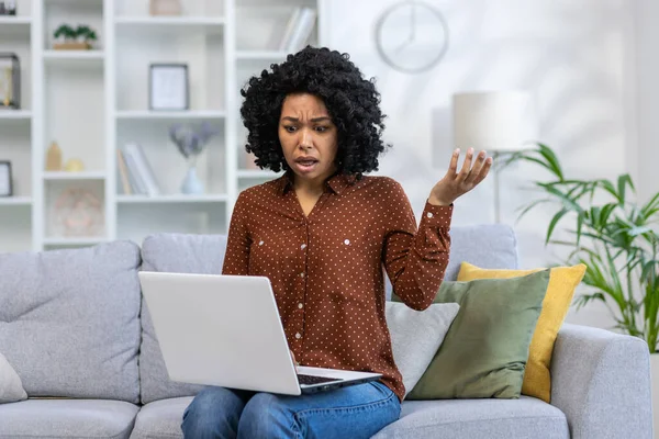 Worried and shocked young African American woman sitting on sofa at home and looking at laptop screen in surprise, spreading arms.