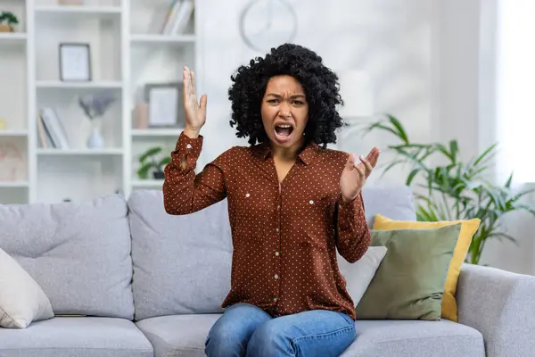 Portrait of a young angry African American woman sitting on the sofa at home, screaming and gesturing emotionally with her hands while looking at the camera.