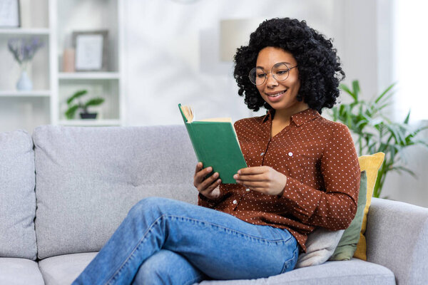 African American young woman relaxing at home, sitting on the sofa and reading a book.