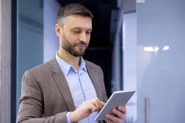 Professional man in smart casual outfit smiling while using a tablet in a modern office setting, depicting work and technology.