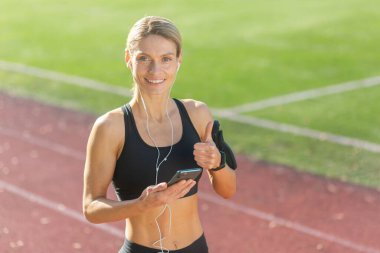 Spor kıyafetli, kulaklıklı, gülümseyen atletik bir kadın telefon kullanıyor ve güneşli bir pistte başparmağını kaldırıyor..