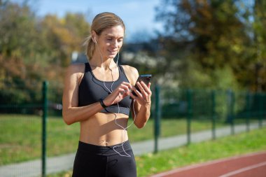 Formda sporcu bir bayan sporcu güneşli bir parkta koşuya çıkmadan önce telefonundan müzik seçer..