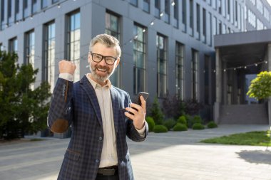 Neşeli bir üst düzey yönetici, modern bir ofisin önünde elinde akıllı bir telefonla zafer jesti yapıyor..