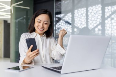 Neşeli genç Asyalı bir iş kadını, ofisinin çalışma yerinde, dizüstü bilgisayarında ve telefonunda yaptığı yumrukla kutlama yaparken başarısını sergiliyor..