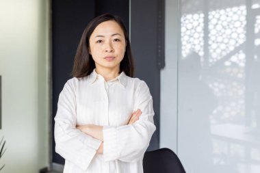 Profesyonel Asyalı iş kadını ofis masasının başında dikilirken kendine güvenerek kollarını kavuşturuyor liderlik ve başarıyı somutlaştırıyor..