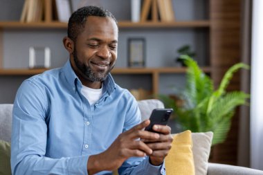 Mutlu Afro-Amerikan adam evinde koltukta oturuyor ve cep telefonuyla gülümsüyor. Yakın plan fotoğraf..