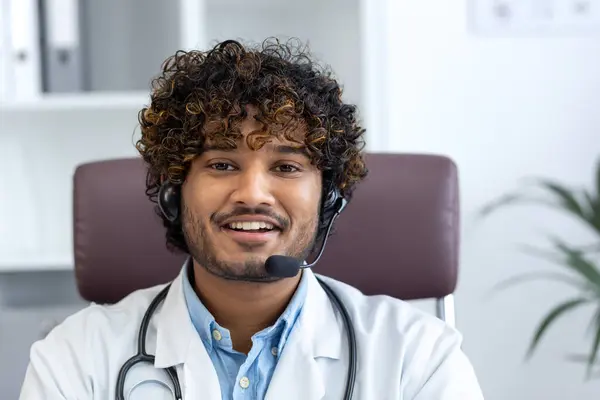 Webcam view, young doctor with headset phone using laptop for video call, doctor cheerfully and friendly consulting patients, smiling and looking at camera, working inside clinic medical room.