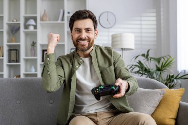 Kanepede oturan gülümseyen adam, rahat bir oturma odasında konsol oyunu oynamaktan zevk alırken muzaffer bir şekilde yumruk tokuşturuyor..