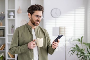 Modern bir ev ortamında elinde kahve fincanı ve akıllı telefonuyla gülen adam rahat ve rahat bir yaşam tarzı yayıyor..