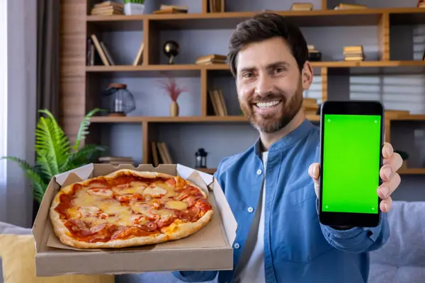 Cheerful adult male presents a mobile phone with a green screen next to a delicious pizza indoors, ready for app promo.