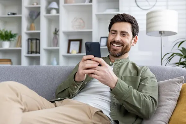 Joyful male in casual attire browsing on mobile phone, exuding relaxation and happiness in a cozy living room.