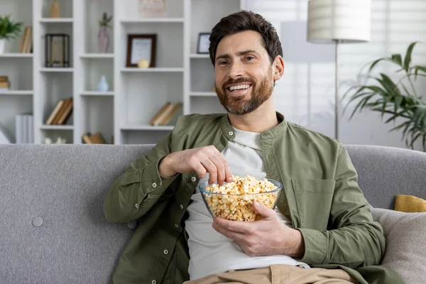 Cheerful adult male relaxes on a couch, holding a bowl of popcorn, a warm and cozy home setting fading into the background.