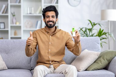 Gülümseyen Hintli adam evdeki kanepede rahatça oturuyor, başparmağıyla işaret ediyor, pozitiflik ve onay gösteriyor..