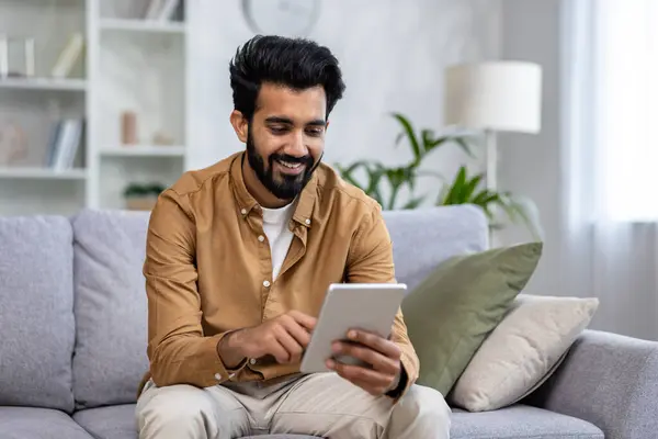 Indian man smiles while browsing on a tablet, comfortably seated on a sofa in a well-lit modern apartment living room.