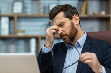İş kıyafetleri içinde ciddi bir olgun adam yorgun hissediyor. Klasik bir ofis ortamında işine odaklanırken gözlerini ovuşturuyor..