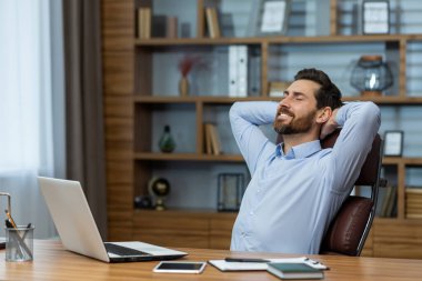 Olgun bir iş adamı iyi organize edilmiş bir ofis ortamında ellerini başının arkasına koyup dinleniyor..