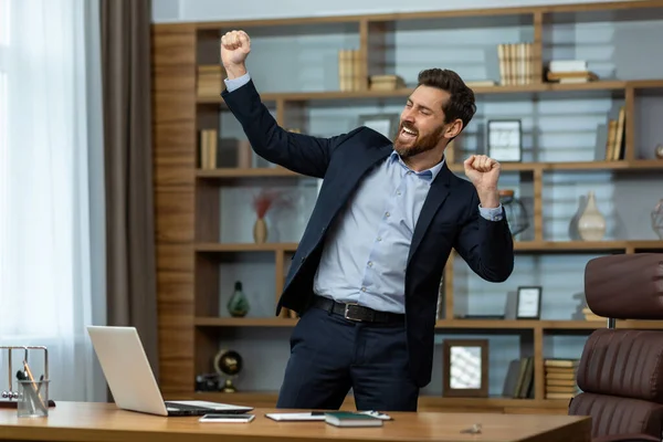 Energetic mature businessman in a smart suit celebrates victory with a fist pump in a well-appointed home office setting.