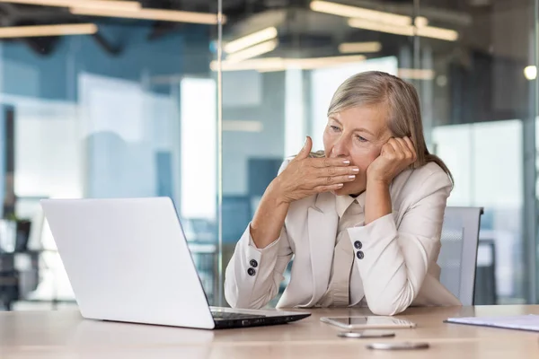 Tired senior woman sitting at desk in front of laptop in office in business suit, head on hand and yawning while covering mouth with hand.