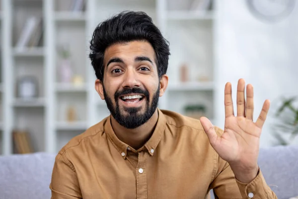 Close-up photo of a young Indian man sitting on the couch at home and chatting online. Says hello and waves at the camera.