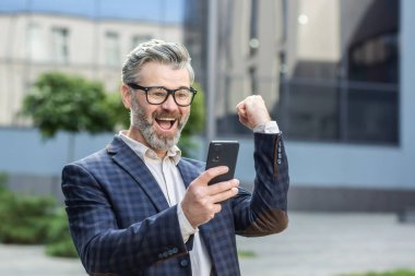 Heyecanlı takım elbiseli son sınıf öğrencisi, arka planda bir ofis binasıyla telefonuyla başarıyı kutluyor..