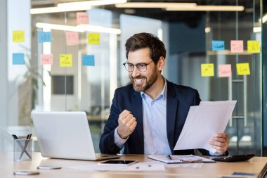 Belgeler, belgeler, raporlar, iş yerinde çalışan başarılı muhasebeci, sonuçlardan memnun olan erkek finansör zaferi kutluyor. Laptop kullanıyor..