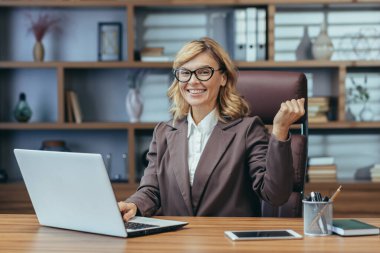 Modern bir ev ofisinde dizüstü bilgisayarla çalışan neşeli, olgun iş kadını kendine güvenini ve profesyonelliğini ifade ediyor..