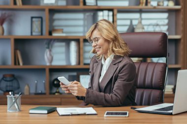 Şık bir ev ofisinde akıllı telefon kullanarak gülümseyen olgun bir iş kadını. Teknoloji ile başarılı kadın profesyonel çoklu görev.