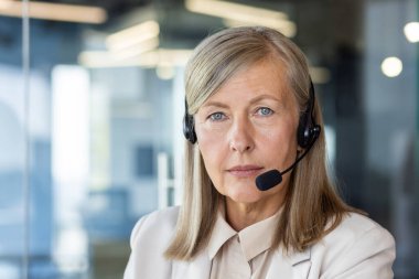 Ofiste oturmuş, ciddi ve kendinden emin bir şekilde kameraya bakan, kulaklık takan yaşlı, gri saçlı bir kadının yakın plan portresi..