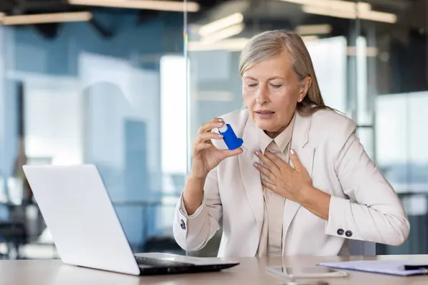 Senior gray-haired businesswoman sitting in the office at the desk and holding her hand to her chest, suffering from asthma and shortness of breath, using an inhaler.