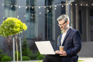 İş elbisesi içinde deneyimli bir finansör. Bir yandan bir dizüstü bilgisayara odaklanırken, diğer yandan da şehir ortamında yapılması gereken kahve işini yönetiyor..