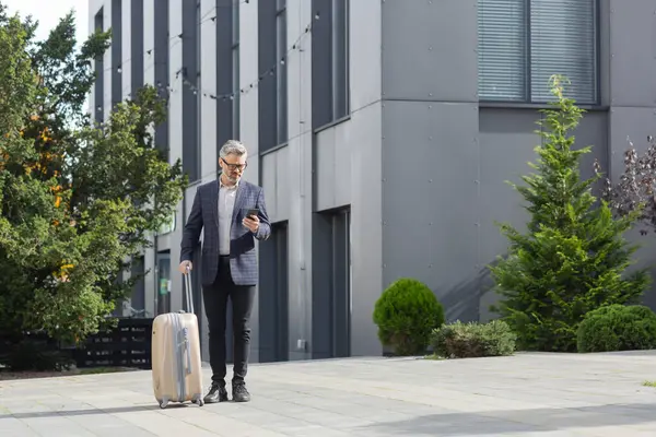 A mature businessman in a suit walks with a suitcase, focused on his phone, by an office building with greenery around.