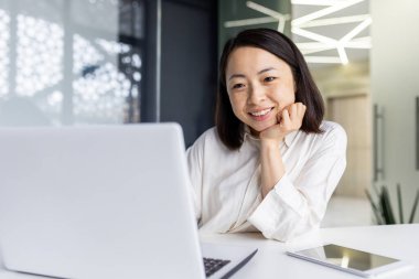 Portatif bilgisayar ve dijital tabletle masaya yaslanırken beyaz bluzlu tatmin olmuş Asyalı kadın çenesini dinlendiriyor. Esmer kadın ofisin içinde gülümseyen dizüstü bilgisayara bakıyor..