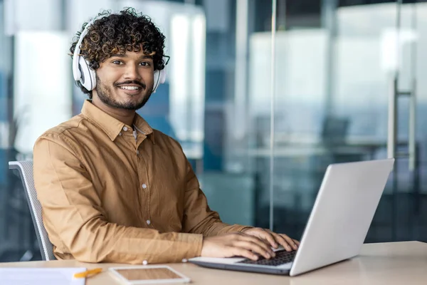 Indian guy with curly hair sitting in wireless headphones at working place with laptop and looking at camera. Confident man listening to playlist for improving focusing on project enhancement indoors.