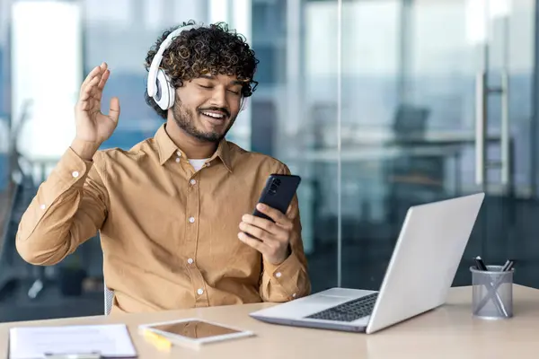 Hindu man in wireless headphones sitting by personal workplace with computer and notepad while looking at phone screen. Relaxed office manager using gadgets for communication with family during break.