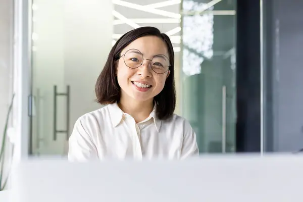 Portrait of pleased asian female in round transparent glasses looking at camera with toothy smile in workspace. Successful project manager with short haircut enjoying productive busy day in office.