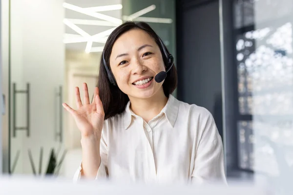 First person view of smiling asian woman in wireless headset with micro looking at camera and making hello gesture. Positive female greeting colleagues while starting online video meeting at office.