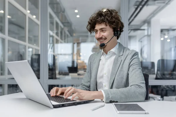 Smiling young businessman with headset working on laptop in modern office, showcasing multitasking and customer service skills. Positive work environment reflected in his cheerful expression.