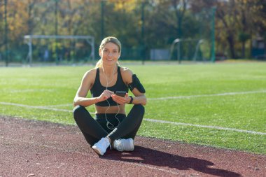 Gülümseyen bir bayan sporcu güneşli bir spor pistinde oturur ve antrenmana ara verdiği sırada akıllı telefonuyla müziğin tadını çıkarır..