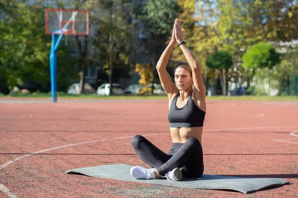 A concentrated young woman sitting on a yoga mat, performing a yoga pose confidently at an outdoor basketball court.