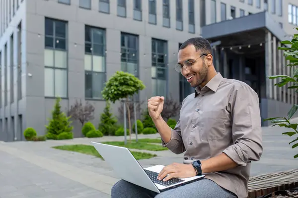 Man celebrating victory successful notification on laptop, businessman holding hand up triumph gesture, working remotely sitting outside office building on bench.