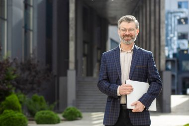 Şık takım elbiseli, elinde kağıtlarla bir ofisin yanında duran gülümseyen olgun bir profesyonel. Tecrübeli liderlik ve finans kavramı.