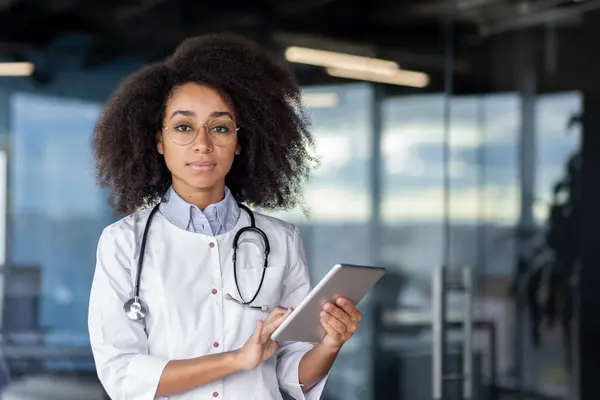 Qualified black woman doctor standing in medical center and holding digital tablet in hands while looking at camera. Serious female physician in uniform ready for meeting patients and giving help.