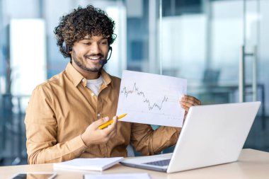 Video konferansını yöneten kablosuz kulaklıklı iyimser İspanyol işveren etkileyici sonuçlarla istatistik çizelgesi gösteriyor. Pazarlama bölümü başkanı başarılı stratejinin sonuçlarını paylaşıyor.