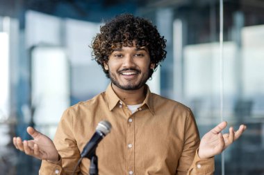 Yakışıklı Arap işadamı şık bir ofiste oturuyor ve mikrofonun önünde konuşuyor. Pozitif erkek işveren iş arkadaşları için kurumsal etkinlik ve el hareketleriyle destekleyici bir konuşma yapıyor..
