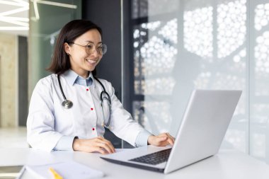 Dizüstü bilgisayarda çalışan ve hastane ofisinde gülümseyen bir kadın doktor. Asyalı sıhhiyeci kadın taşınabilir bilgisayarda online danışmanlık yapıyor ve iyi haberler aldıktan sonra mutlu oluyor..