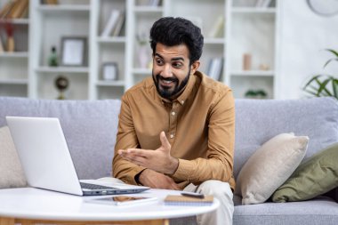 Genç Hintli adam internette konuşuyor, evdeki kanepede dizüstü bilgisayarın önünde oturuyor, elleriyle kameraya gülümsüyor..