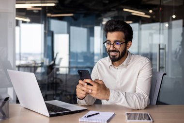Genç Hintli bir adam bir masada oturmuş gülümsüyor ve cep telefonu kullanıyor..